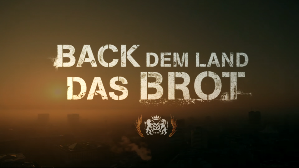 Motivbild: Back dem Land das Brot. Schriftzug mit dem Bäckerwappen im Morgenhimmel mit aufgehender Sonne über einer Stadt.