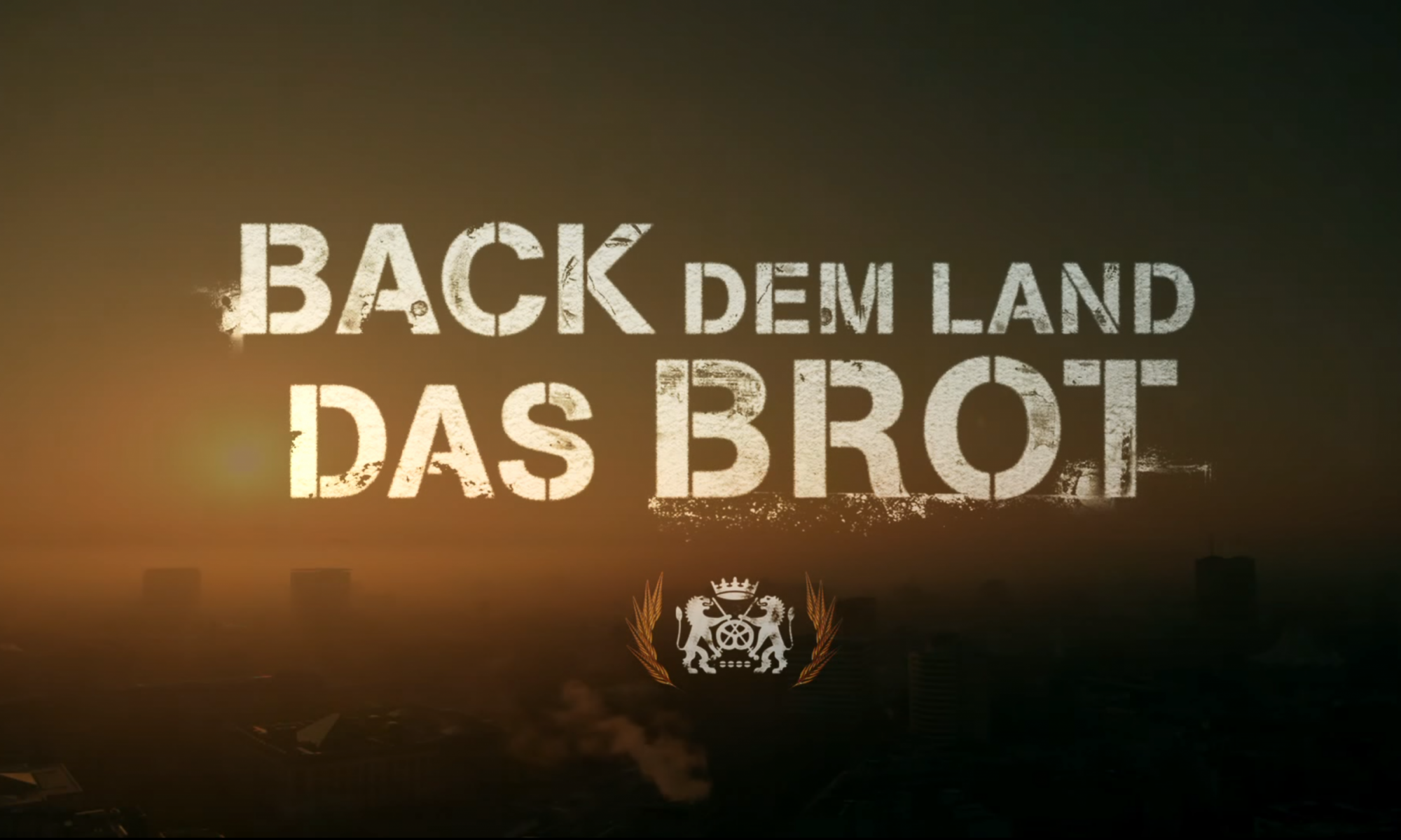 Motivbild: Back dem Land das Brot. Schriftzug mit dem Bäckerwappen im Morgenhimmel mit aufgehender Sonne über einer Stadt.